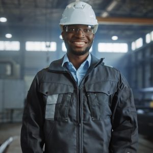 Happy Professional Heavy Industry Engineer/Worker Wearing Uniform, Glasses and Hard Hat in a Steel Factory. Smiling African American Industrial Specialist Standing in a Metal Construction Manufacture.