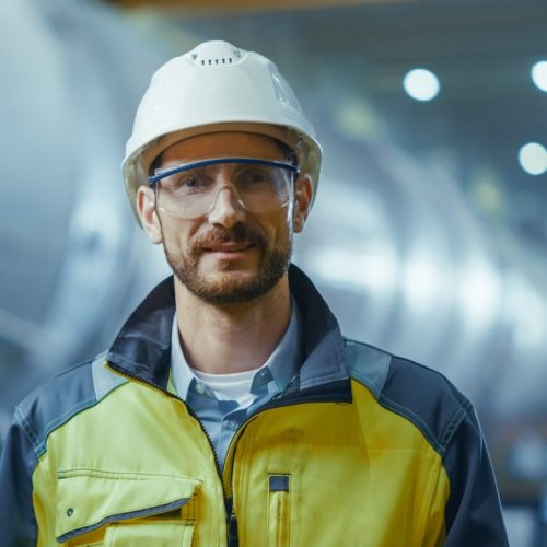 Portrait of Smiling Professional Heavy Industry Engineer / Worker Wearing Safety Uniform, Goggles and Hard Hat. In the Background Unfocused Large Industrial Factory where Welding Sparks Flying